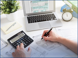 person looking at tax forms and a calculator in front of a laptop