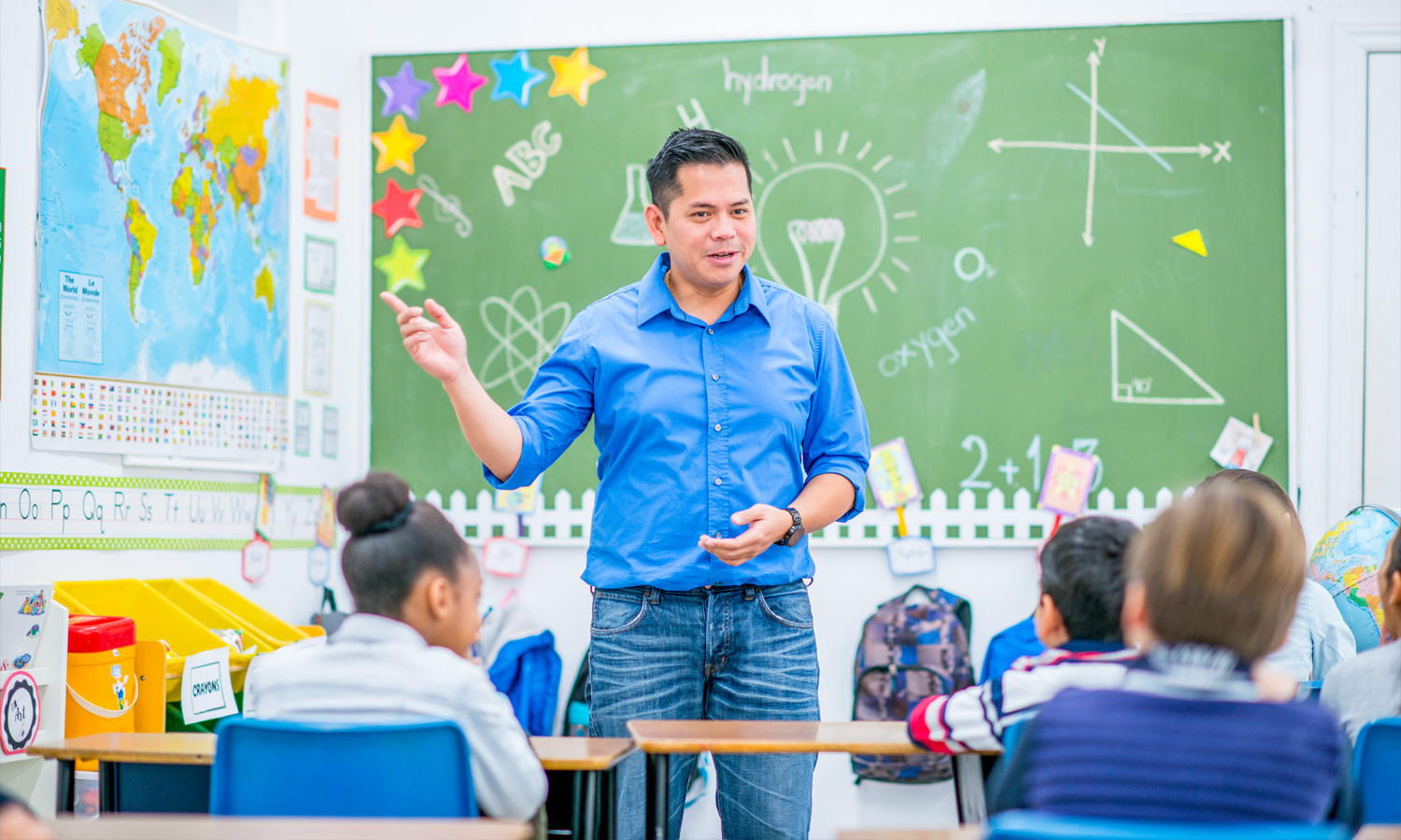 man standing in front of a green chalkboard talking to children sitting in desks